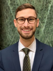 Headshot of Ryan J. Specht-Boardman, wearing glasses, a blue blazer, and green patterned tie.
