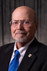 A man "Ray Schroeder" is dressed in a suit with a blue tie and wearing glasses.