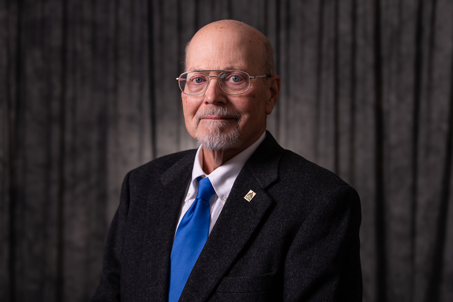A man (Ray Schroeder) is dressed in a suit with a blue tie and wearing glasses.