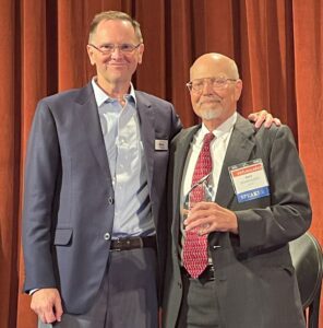 Two men (Bob Hansen and Ray Schroeder) stand on stage, smiling at the camera. Ray holds an award.