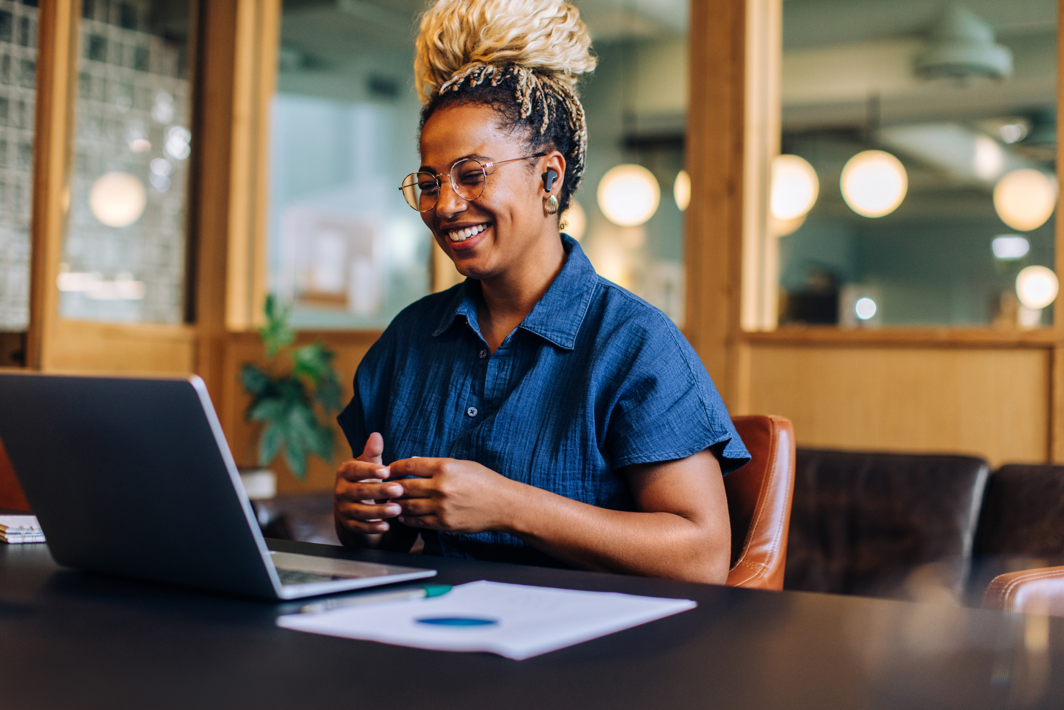 A cheerful woman engaging in a video conference in a contemporary office environment using a laptop.