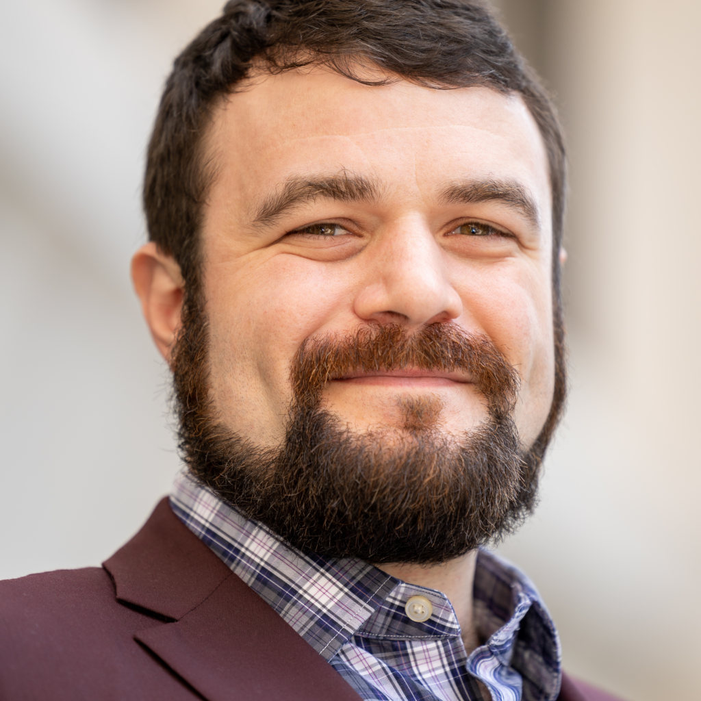 A man (Jordan DiMaggio) with a beard is dressed in a suit with a plaid dress shirt for a headshot.