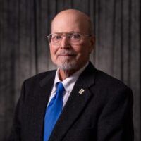 A man (Ray Schroeder) is dressed in a suit with a blue tie and wearing glasses.